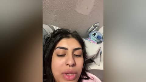 Media: Video of a young woman with medium brown skin, dark hair, and a septum piercing, lying on a bed with a stuffed Stitch toy.