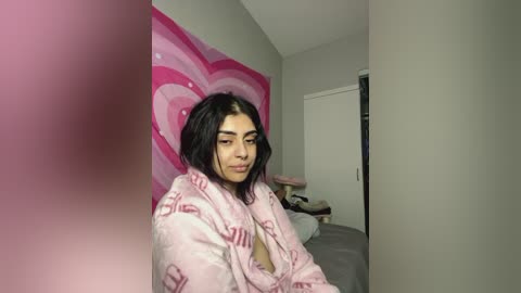 Media: Video of a young woman with medium skin tone, dark hair, and light makeup, wearing a pink, fluffy robe with text, standing in a minimalist, beige-walled bedroom with a large, pink, heart-shaped wall art.