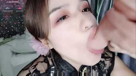 Media: A close-up video of an East Asian woman with fair skin, pink eyeshadow, and dark hair, wearing a black lace dress, performing oral sex on a circumcised penis.