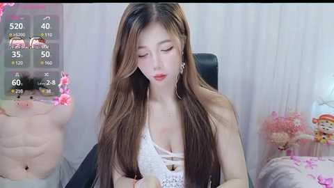 Media: A video of an East Asian woman with long, straight brown hair, wearing a white crochet top, sitting on a black chair, surrounded by pink flowers and a teddy bear.