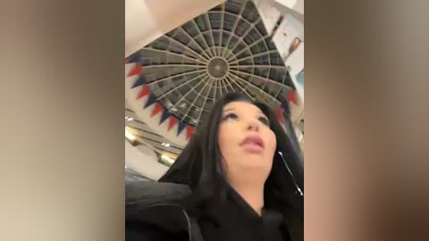 Media: Video of a woman with long black hair, wearing a black coat, standing under a circular ceiling with a star pattern and red, blue, and gray accents.