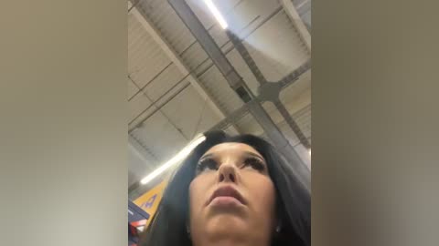 Media: A low-angle video of a woman with long black hair and light skin, wearing dark lipstick, looking upward. The background features industrial ceiling lights and pipes, suggesting an indoor setting.