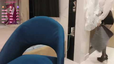 Media: Video of a modern interior with a blue upholstered chair, a white-clad woman holding a grey garment, and a digital screen displaying colorful text in the background.