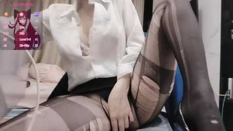 Media: Video of a woman in a white blouse, black skirt, and sheer stockings, sitting on a bed with a computer monitor displaying a video feed.