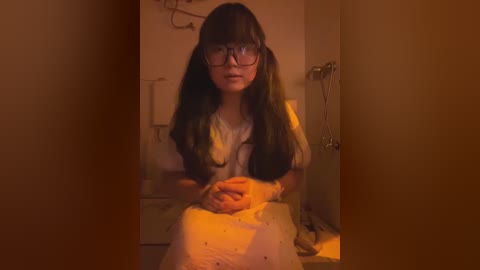 Media: Video of a young Asian woman with long black hair, wearing glasses, a white shirt, and a pink dress, seated in a dimly lit room with a phone charger hanging from the wall.