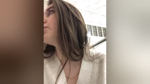 Media: Video of a young woman with long, wavy brown hair, wearing a white sweater, standing indoors with a blurred background showing a ceiling and window.