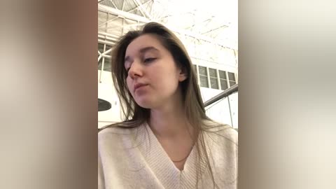 Media: A video of a young woman with light skin, brown hair, and a neutral expression, wearing a white cardigan, standing in an airport with modern architecture and large windows in the background.