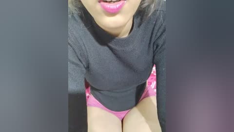 Media: Video of a woman in a dark gray sweater and pink shorts, with light skin and pink lipstick, sitting against a dark background.