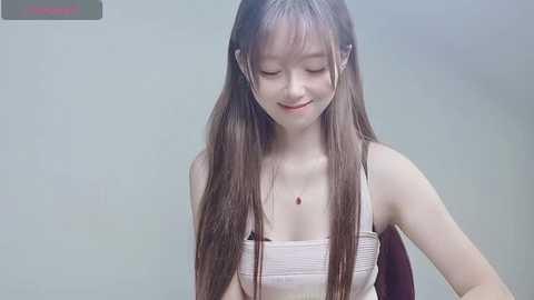 Media: Video of a smiling East Asian woman with long, straight brown hair, wearing a white tank top, sitting against a plain light gray wall.