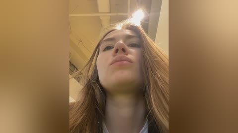 Media: A video of a young woman with long, light brown hair, fair skin, and light makeup, captured from a low angle, looking directly into the camera. She appears to be indoors with a warm, diffused lighting.