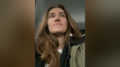 Media: Video of a young woman with wavy, light brown hair and fair skin, wearing a dark jacket, looking upwards with a thoughtful expression. Background is blurred, possibly indoors.