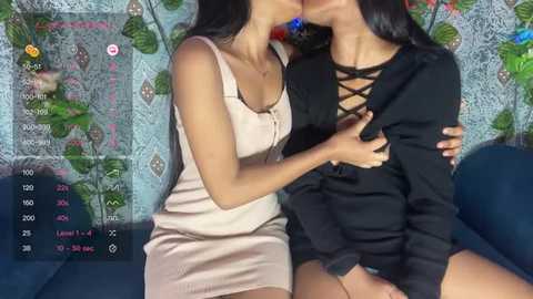 Media: Video of two women in a passionate kiss, one in a beige dress, the other in a black lace top. Background features a blue sofa and a colorful, floral-patterned wall.