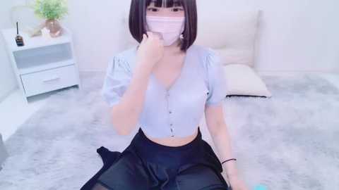 Media: A video of an Asian woman with straight, black hair and a pale complexion, wearing a light blue V-neck top, black skirt, and a pink face mask, kneeling on a plush, grey carpet in a bright, minimalistic room.