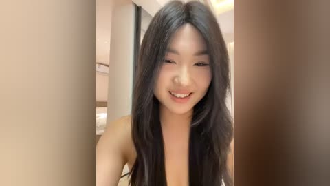 Media: Video of a young Asian woman with long, straight black hair, smiling, wearing a sleeveless top. She stands in a modern, brightly lit room with beige walls and a minimalistic design.