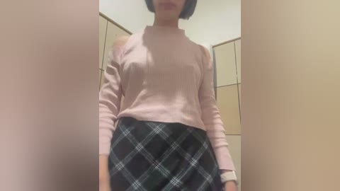 Media: Video of a woman with short black hair wearing a light pink turtleneck sweater and a black plaid skirt, standing in a beige-walled room with a door and cabinets in the background.