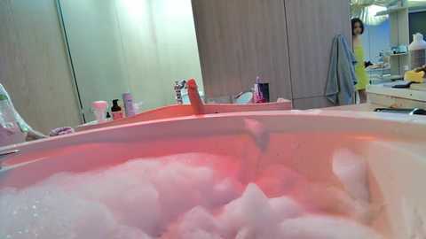 Media: Video of a bathtub filled with pink bubble bath, reflecting a person in a yellow shirt standing in a modern bathroom with beige walls, shelves, and a mirror.