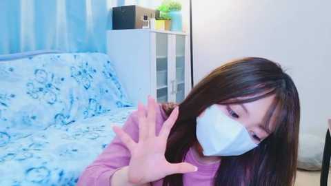 Media: Video of an East Asian woman with long black hair, wearing a white mask, pink top, and blue patterned blanket, gesturing with her hands in a modern, brightly lit bedroom.