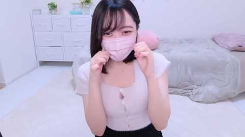 Media: Video of an East Asian woman with straight black hair, wearing a pink mask, white top, and black pants, standing in a minimalist, white bedroom with a bed, dresser, and potted plants.