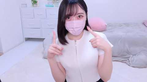 Media: Video of an East Asian woman with straight black hair and light skin, wearing a pink face mask, white zip-up top, and giving a thumbs-up sign in a minimalist, white bedroom with a bed, dresser, and plants.