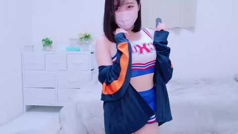 Media: Video of an East Asian woman in a white face mask, blue cheerleader outfit, and black jacket, standing in a minimalist white bedroom with drawers and a bed.