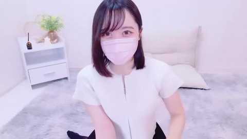 Media: A video of an Asian woman with shoulder-length black hair, wearing a white top, pink face mask, and black pants, kneeling on a soft gray carpet in a minimalist, white-walled room.