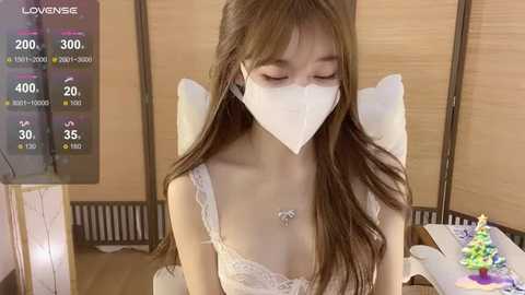 Media: A video of an Asian woman with long brown hair, wearing a white face mask, white lace bra, and sitting on a chair in a cozy room with a temperature display.