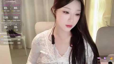 Media: Video of an East Asian woman with long black hair, wearing a white lace top, sitting indoors. The background shows beige curtains and a white chair.