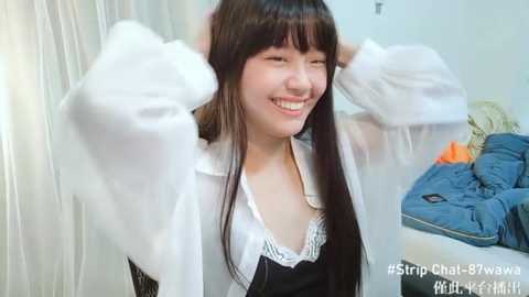 Media: Video of a young East Asian woman with straight black hair, wearing a white blouse and black vest, smiling while adjusting her hair. Background features a blue couch with a blanket and a white curtain.