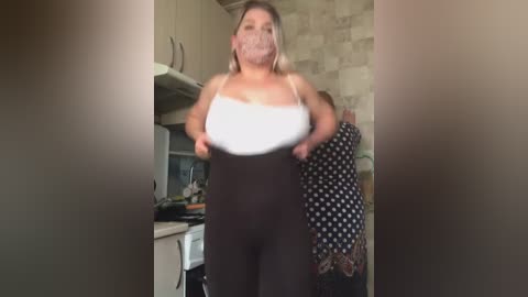 Media: A video of a plus-sized woman in a white tank top and black polka-dot leggings in a kitchen with beige tiled walls.