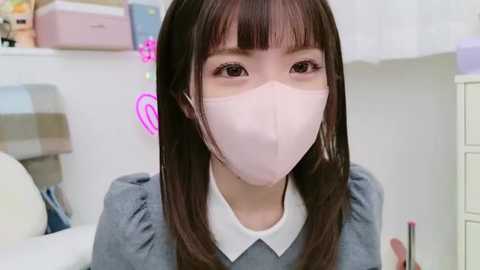 Media: Video of an Asian woman with long brown hair, wearing a light pink face mask, grey sweater, and white collar. Background features a white room with pink neon lights, a striped pillow, and a white desk.