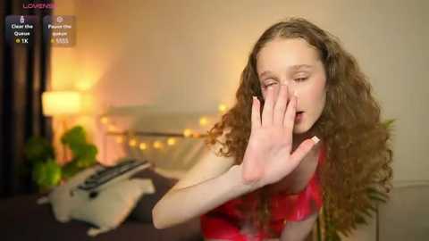 Media: Video of a young woman with long, curly brown hair, wearing a red lace top, covering her face in distress. Background features a bed with a white lamp and a green plant, warm ambient lighting.