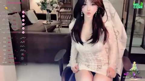 Media: A video of an Asian woman with long black hair and fair skin, wearing a white lace dress, sitting in a modern living room with a sofa and bookshelves.