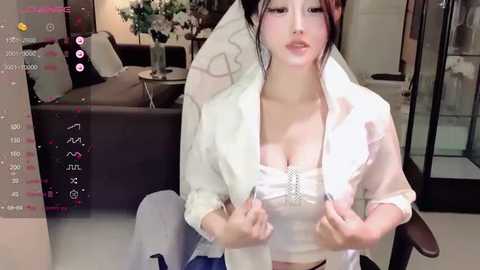 Media: Video of an Asian woman with pale skin, wearing a white blouse and jacket, sitting in a modern living room with a black couch and flowers in the background.