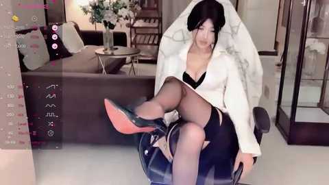 Media: Video of a young Asian woman with black hair, dressed in a white jacket, black bra, and skirt, sitting on a glass chair, surrounded by modern furniture in a well-lit room.