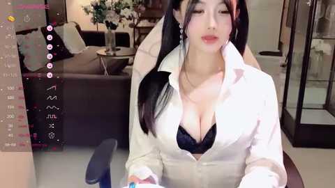 Media: A video of an East Asian woman with long black hair, wearing a white button-up shirt and black bra, seated in a modern living room with a brown couch and flowers.