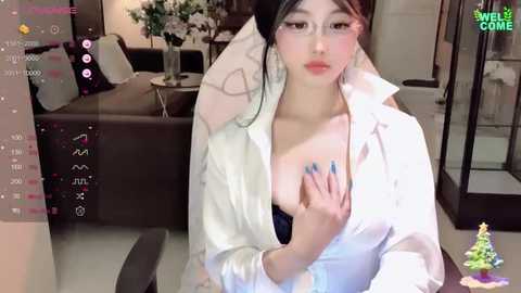 Media: Video of an Asian woman with fair skin and black hair, wearing a white shirt, revealing a black bra, holding a phone, in a modern living room with flowers.