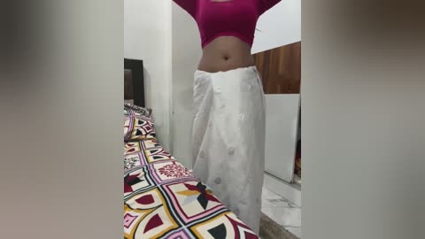 Media: A video captures a woman in a bright pink crop top and white dhoti, standing in a narrow, dimly lit hallway with patterned bedsheets and wooden flooring.