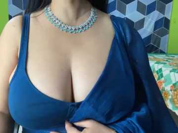 Media: Video of a woman with large breasts in a low-cut, deep blue velvet dress, adorned with a sparkling necklace. Background features a geometric patterned wall and colorful bedspread.