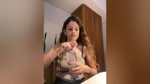 Media: Video of a young woman with long, wavy brown hair, wearing a beige tank top and black pants, filling a water bottle in a modern, minimalist room with wooden paneling and green potted plants.