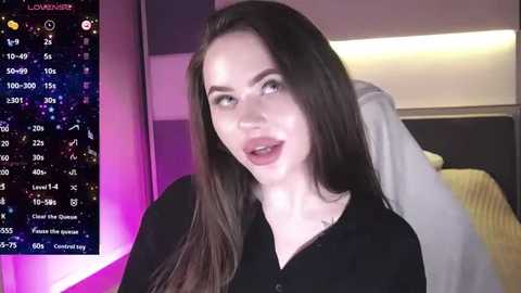 Media: Video of a smiling, fair-skinned woman with long brown hair, wearing a black blouse, standing in a modern bedroom with purple and white lighting.