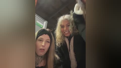 Media: Video of two smiling women with curly blonde and straight blonde hair, wearing black coats, standing inside a dimly lit room.