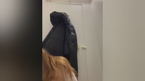 Media: Video of a person with long blonde hair, wearing a black puffy jacket, walking into a dimly lit hallway with white walls. The person's face is not visible.