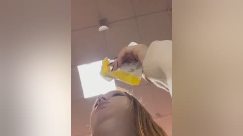 Media: Video of a young woman with fair skin and light brown hair, wearing a white sweater, cleaning a ceiling light with a yellow sponge. The room has beige walls and a fluorescent light.