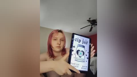 Media: A video of a young, fair-skinned person with shoulder-length pink hair, topless, holding a tablet displaying a \"tip jar\" prompt. The background includes a ceiling fan and a red wall.