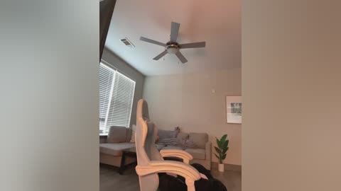 Media: Video of a modern, minimalist living room with beige walls, a gray couch, and a wooden office chair. A ceiling fan and window blinds are visible.