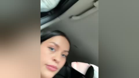 Media: Video of a young woman with fair skin, blue eyes, and straight black hair, wearing a black jacket and white shirt, seated in a car, looking relaxed.