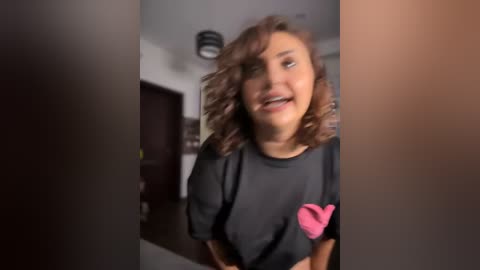 Media: A video of a young woman with medium brown skin and shoulder-length wavy hair, wearing a black t-shirt with a pink heart graphic, smiling in a dimly lit room with dark wooden furniture.