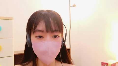 Media: A video of an East Asian woman with long black hair, wearing a pink face mask, black headphones, and a sleeveless top, indoors with a white wall, yellow light, and a white cabinet.