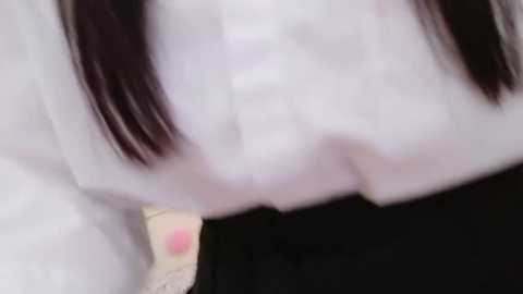 Media: A blurry video showing a person's upper body in a white shirt and black skirt, with long black hair partially obscuring the face. The background is out of focus.