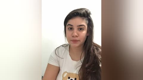 Media: Video of a young woman with long, dark hair, fair skin, and brown eyes, wearing a white T-shirt with a teddy bear graphic. She has a neutral expression against a plain white and brown background.